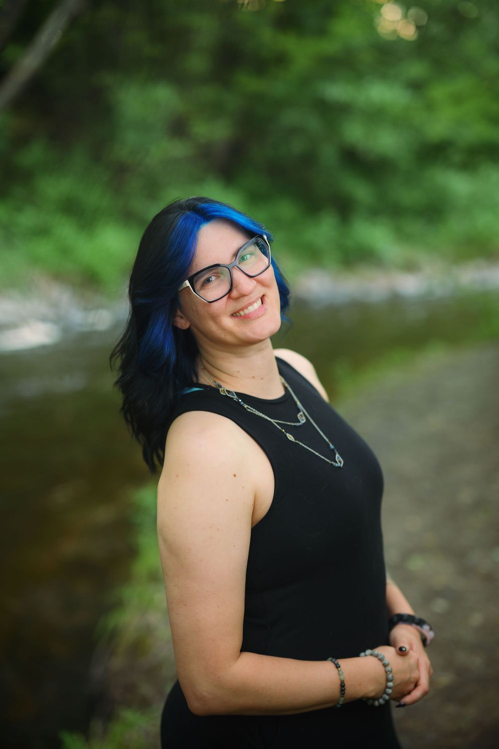 Smiling woman with blue hair and glasses wearing a black top, posing outdoors near a creek surrounded by green trees.