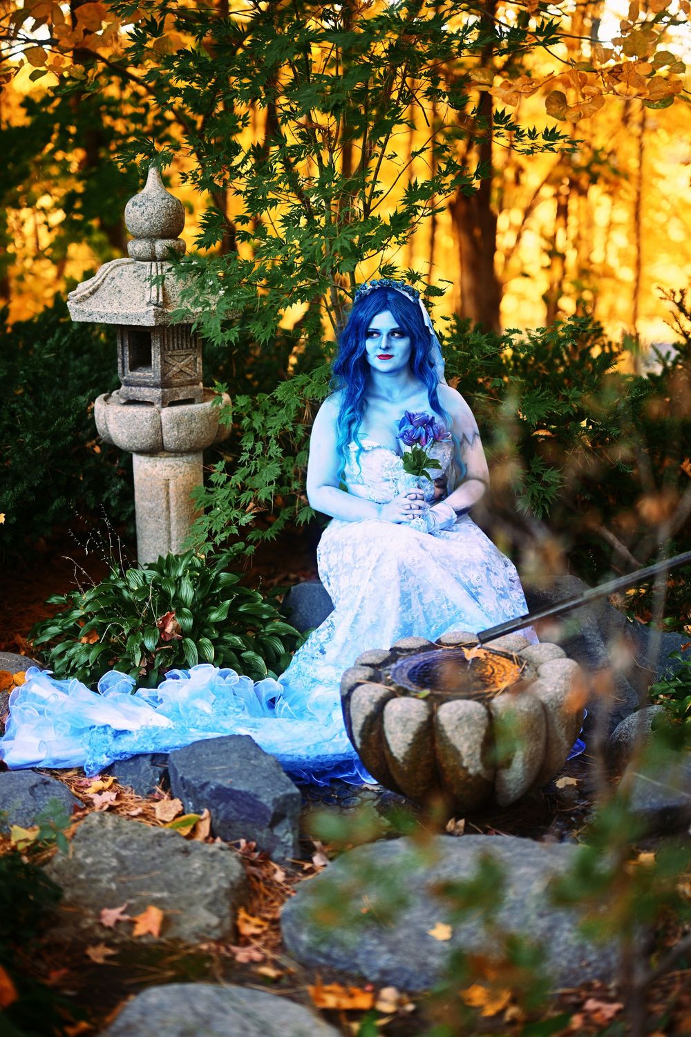 Woman in blue corpse bride costume sits in autumn garden beside stone lantern, holding a blue rose.