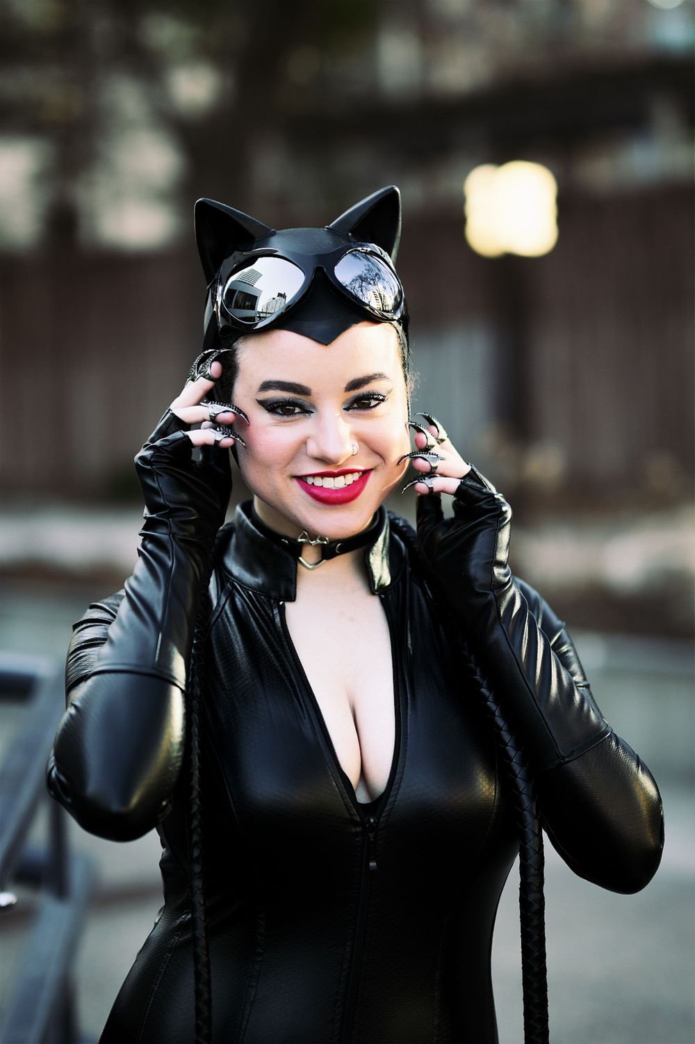 Woman in black latex Catwoman costume with cat ears, goggles, and whip, smiling outdoors at a cosplay event.