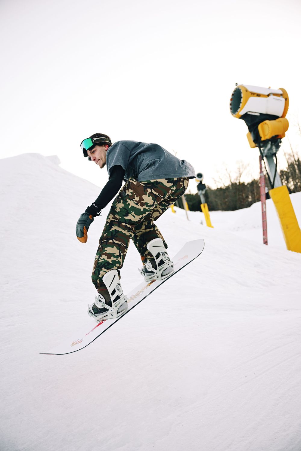 Snowboarder in camo pants and gray jacket performing a jump trick on a snowy ski slope near a snow cannon.