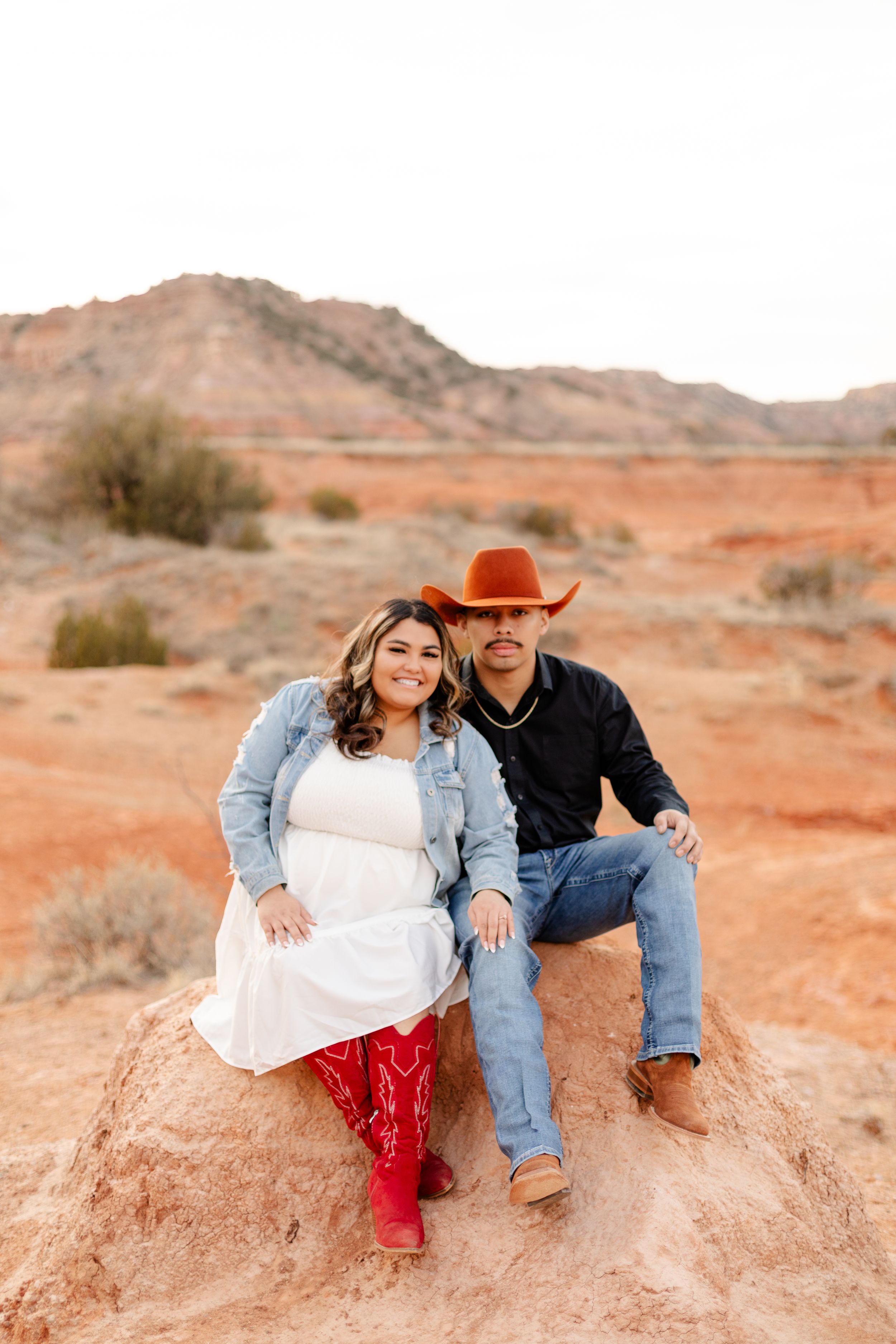 Desert engagement photo with couple sitting on red rock formation at sunset.