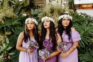 Three bridesmaids in purple dresses wearing floral crowns hold bouquets in a tropical setting.