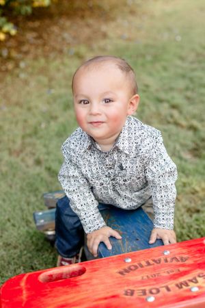A toddler in a patterned shirt smiles while crawling near a red surface outdoors.