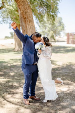 Newlyweds celebrate with joy under a tree, groom raising his arm in triumph.