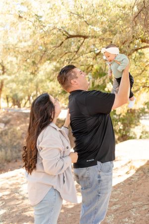 Parents playfully lift their baby in the air during a sunny outdoor photo session.