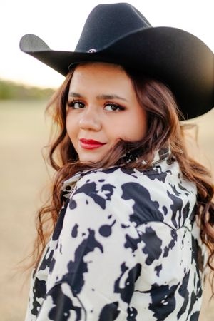 A cowgirl in a black western hat and cow print shirt smiles at the camera in an outdoor setting at sunset.