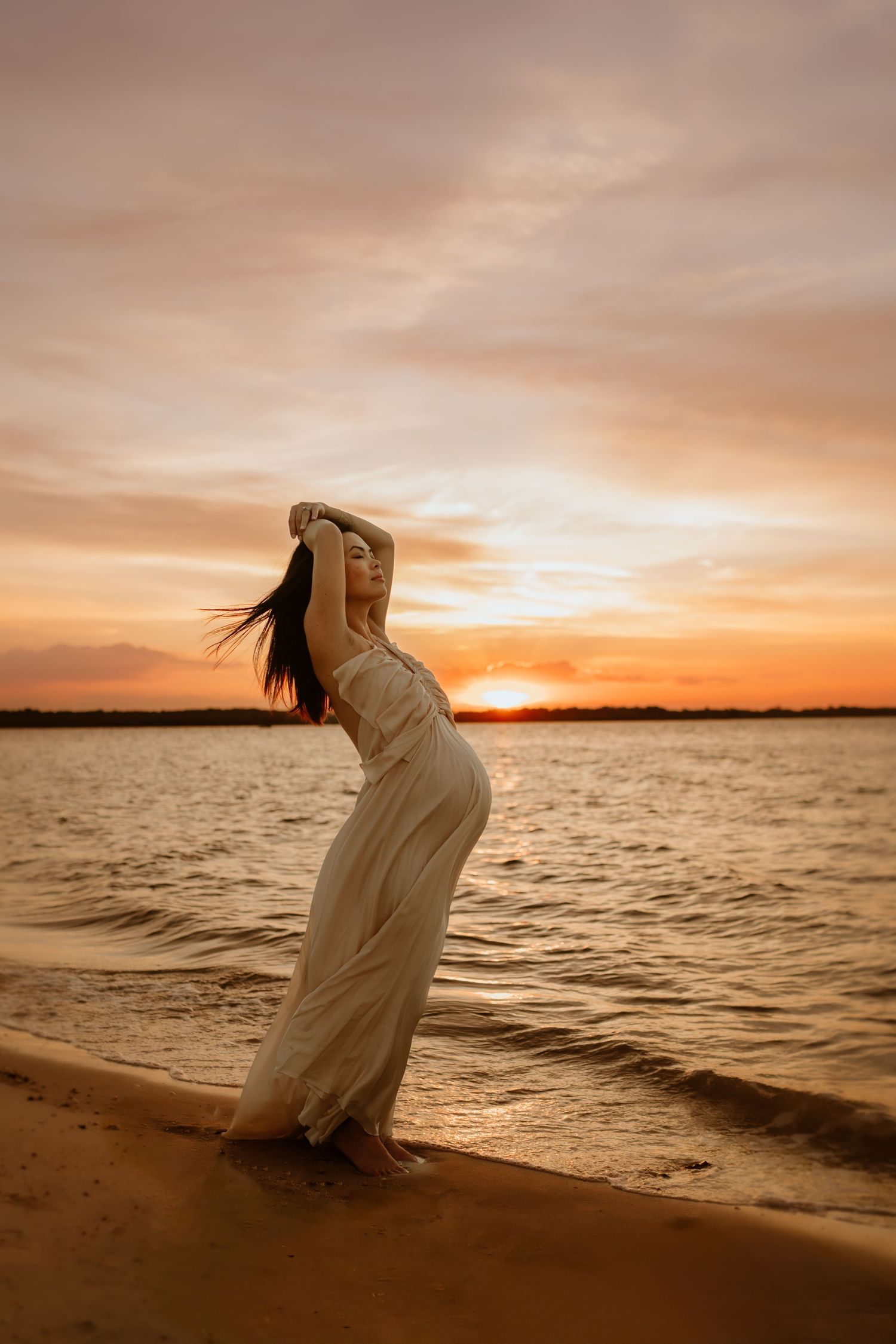 Silhouetted figure poses on a driftwood branch at sunset on a beach with golden light reflecting on the water.