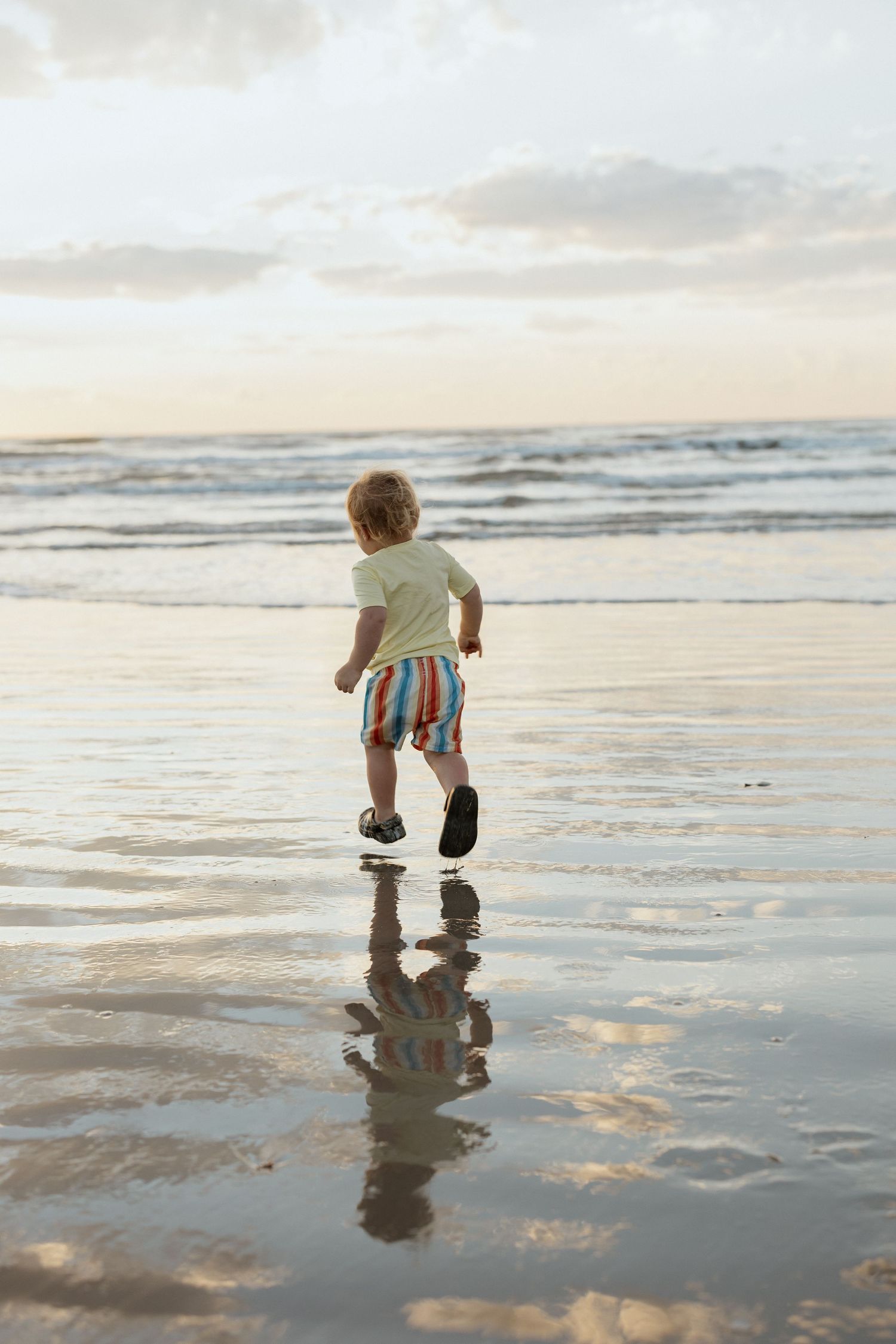 Small child running on wet sand at beach during sunset with reflection in shallow water.