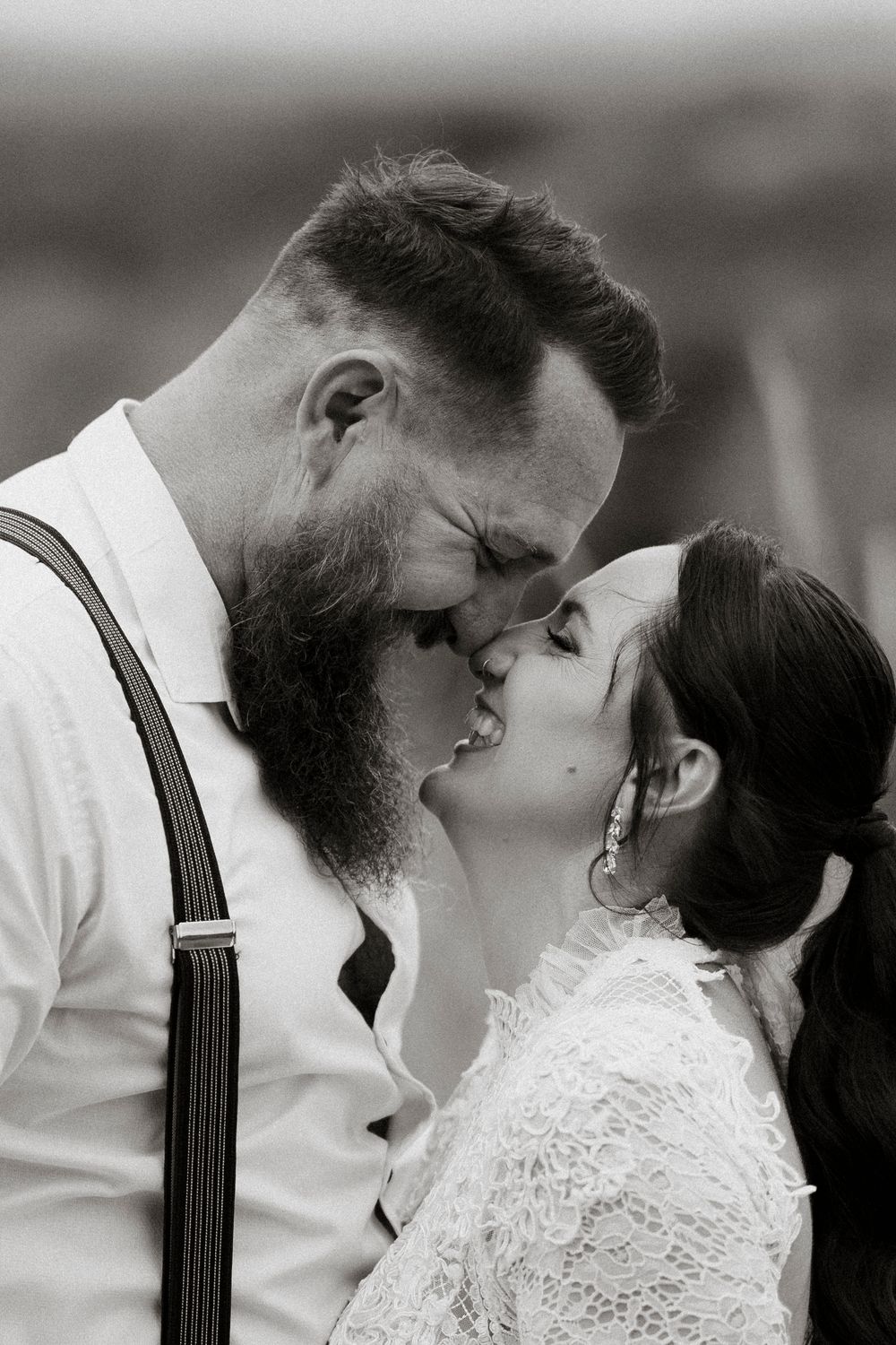 Black and white photo of a bearded groom in suspenders kissing his bride in lace dress at their wedding.