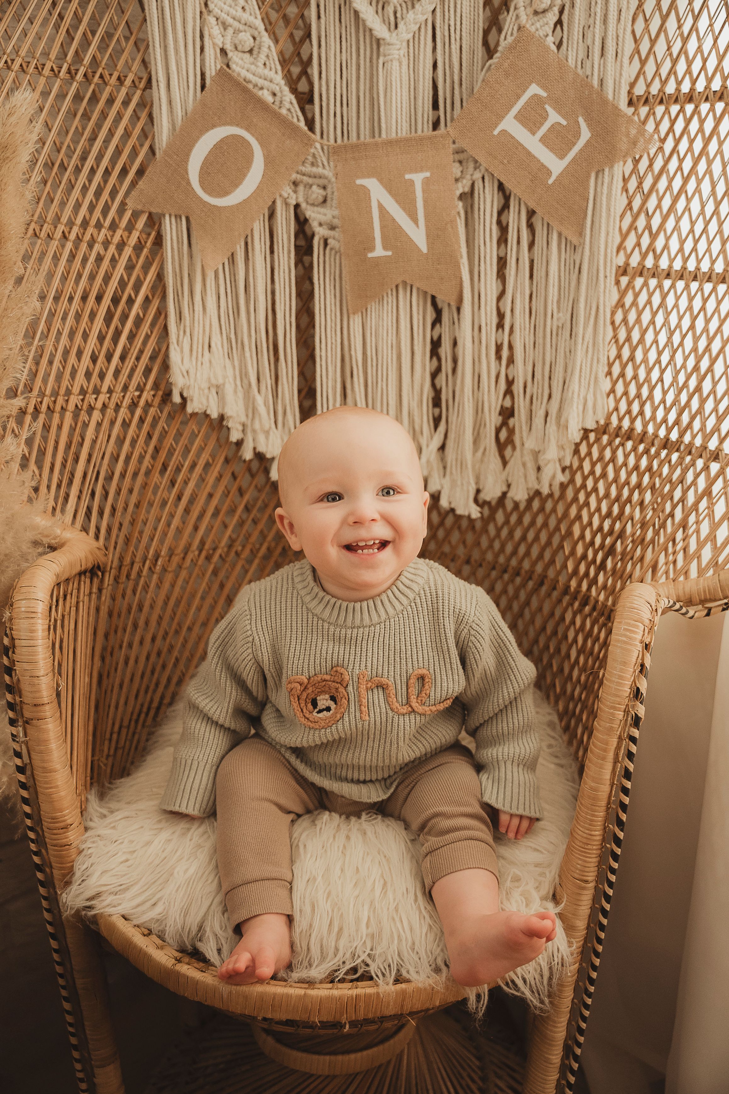 Smiling baby sits in rattan peacock chair wearing beige knit sweater with teddy bear design under ONE burlap banner.