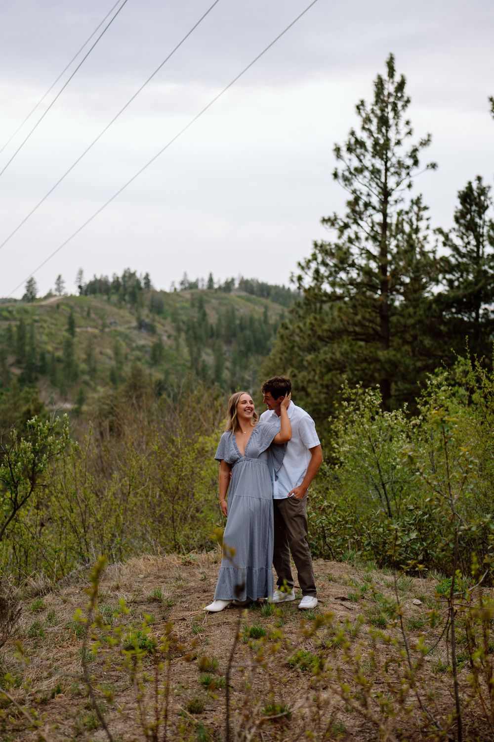A couple stands together on a hillside trail surrounded by pine trees and power lines against an overcast sky.