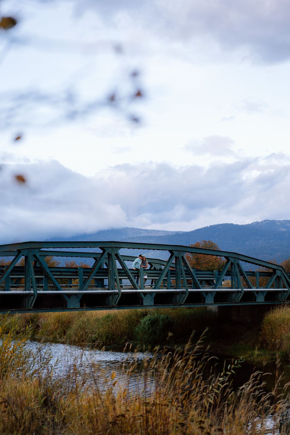 A rustic green metal bridge spans across a serene lake with mountains and cloudy skies in the background.
