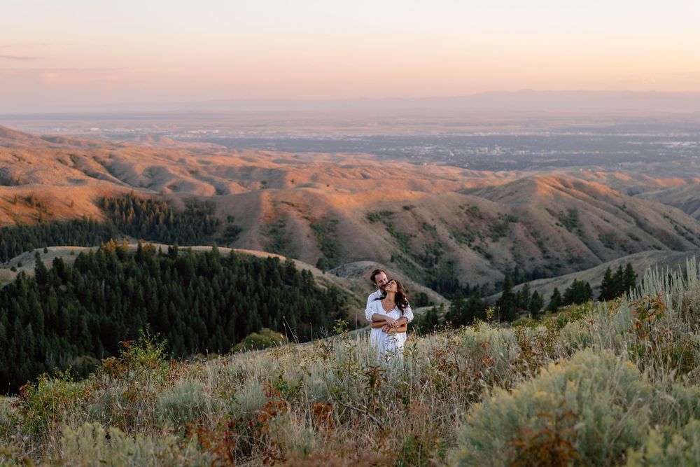 A couple shares a romantic moment at sunset on a scenic mountain overlook surrounded by rolling hills and sagebrush.