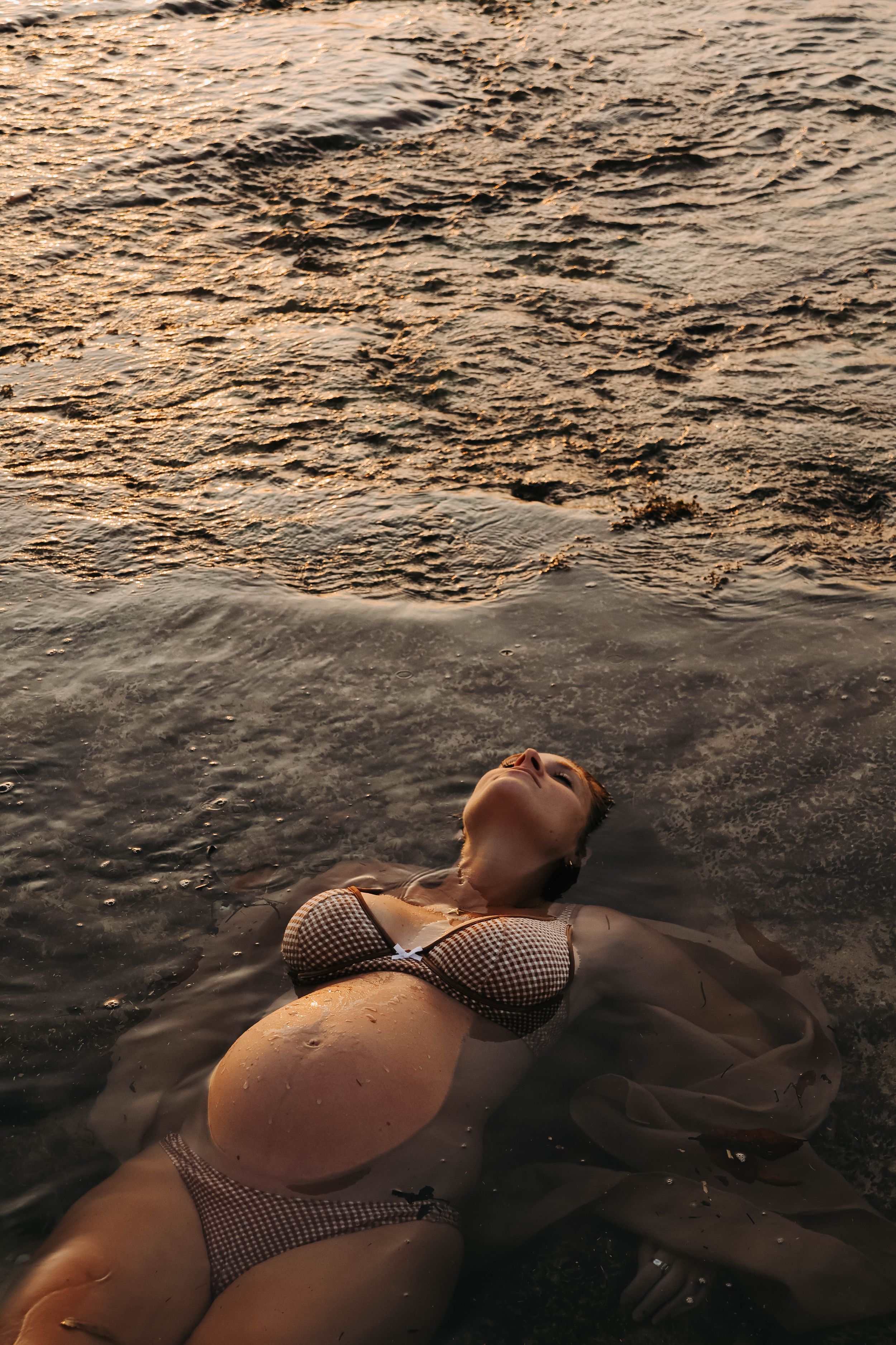 Pregnant woman in bikini lying in shallow ocean waves at golden sunset.