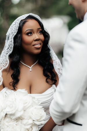 Bride with radiant smile and soft makeup, looking lovingly at her partner in an outdoor wedding setting.