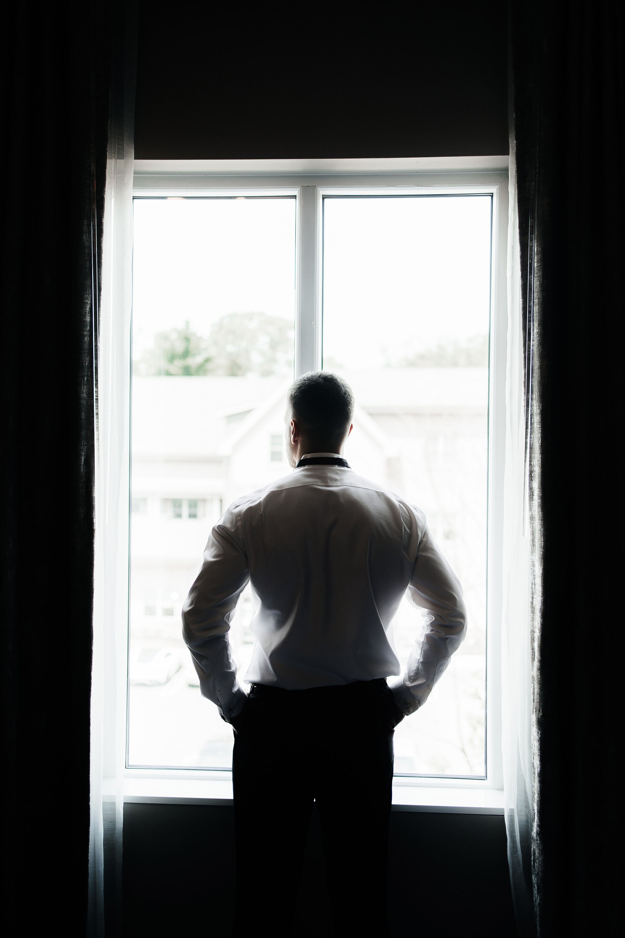 A silhouetted of a groom stands at a window in a dark room, creating a dramatic backlit effect.