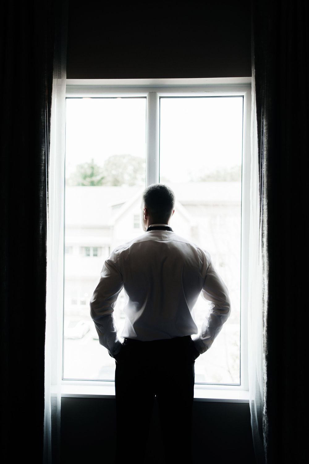 A silhouetted of a groom stands at a window in a dark room, creating a dramatic backlit effect.