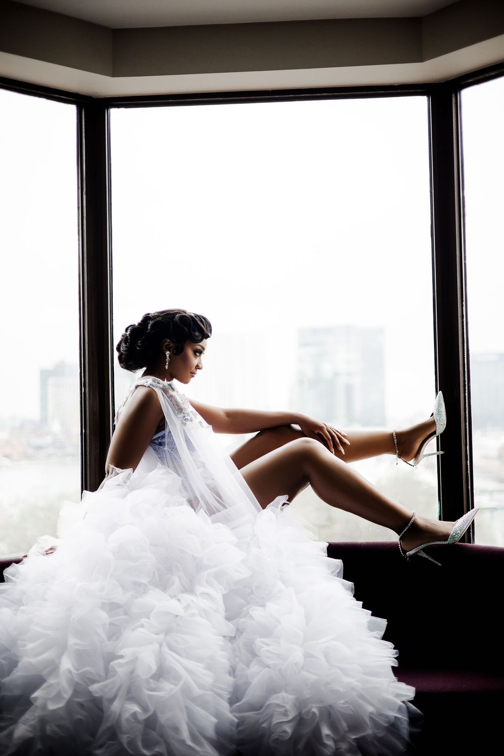 A bride in a white ruffled wedding dress reclines on a couch near large windows with a city view.