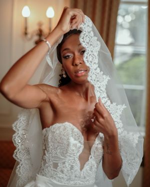 A bride wearing an elegant white lace wedding dress with a delicate veil poses near a window in warm lighting.