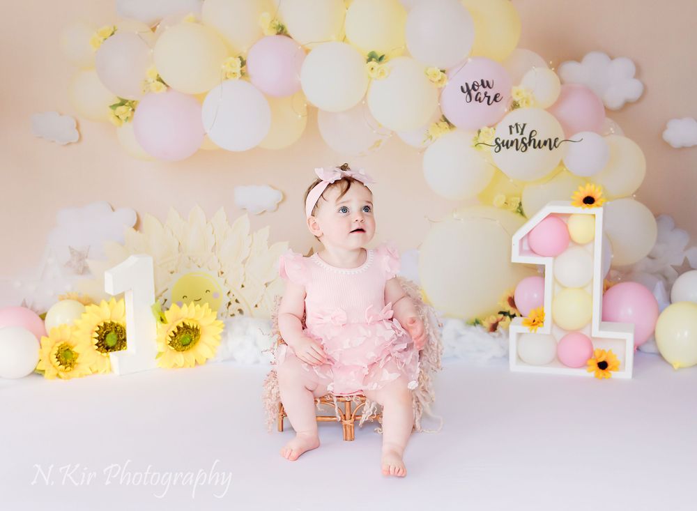 Child is happy and smiling sitting on a chair at her first birthday cake smash session. The scene is bring and colourful. With balloons.