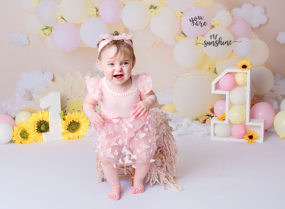 Child is happy and smiling sitting on a chair at her first birthday cake smash session. The scene is bring and colourful. With balloons.