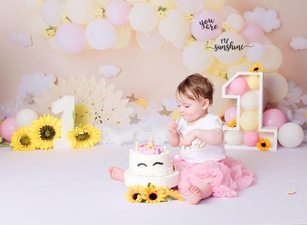 Child eats her cake at her 1st birthday cake smash session. The scene is colourful with sunflowers and a pink tulle dress.