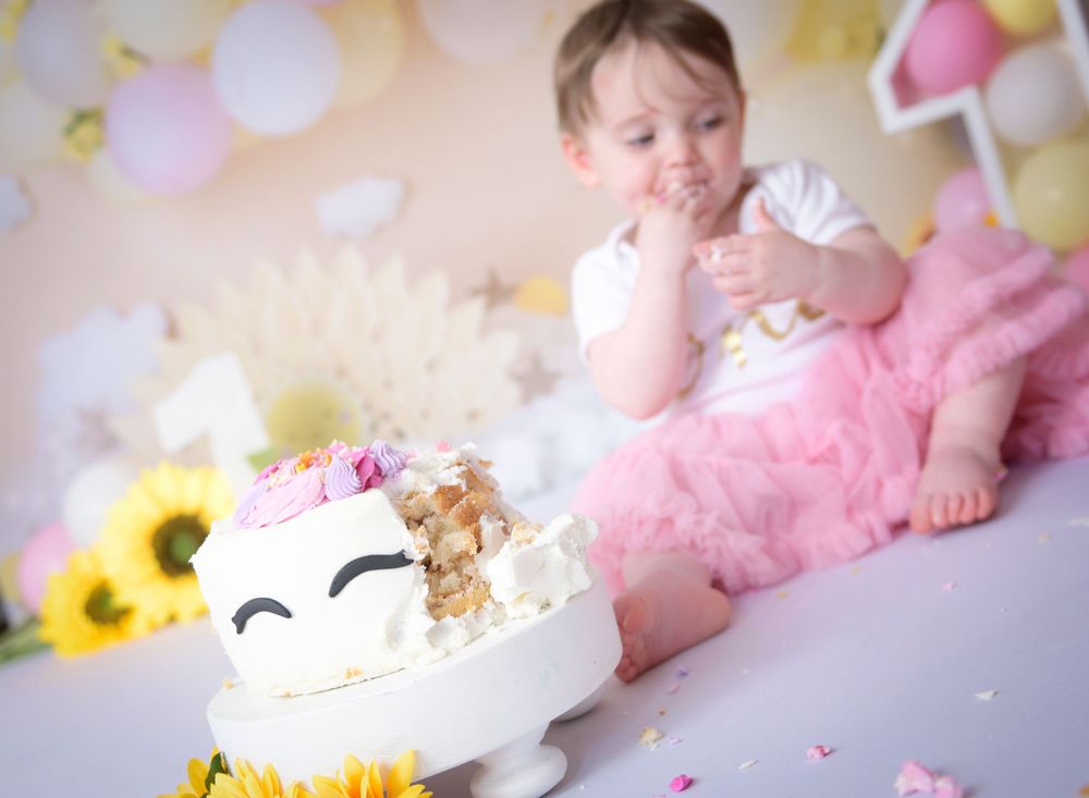 Child eats her cake at her 1st birthday cake smash session. The scene is colourful with sunflowers and a pink tulle dress.