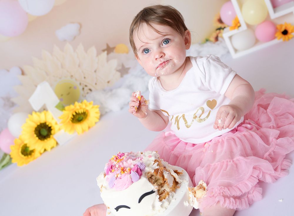 Child eats her cake at her 1st birthday cake smash session. The scene is colourful with sunflowers and a pink tulle dress.