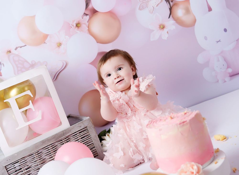 Little girls claps her hands at the cake smash session. Pastel pinks and yummy squidgy cake with lots of balloons and bunnies.