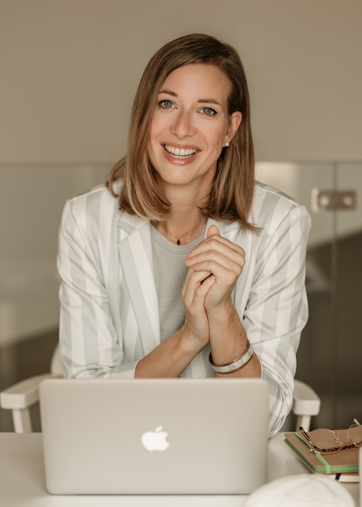 Professional woman in white blazer smiling while working on laptop in modern office setting.
