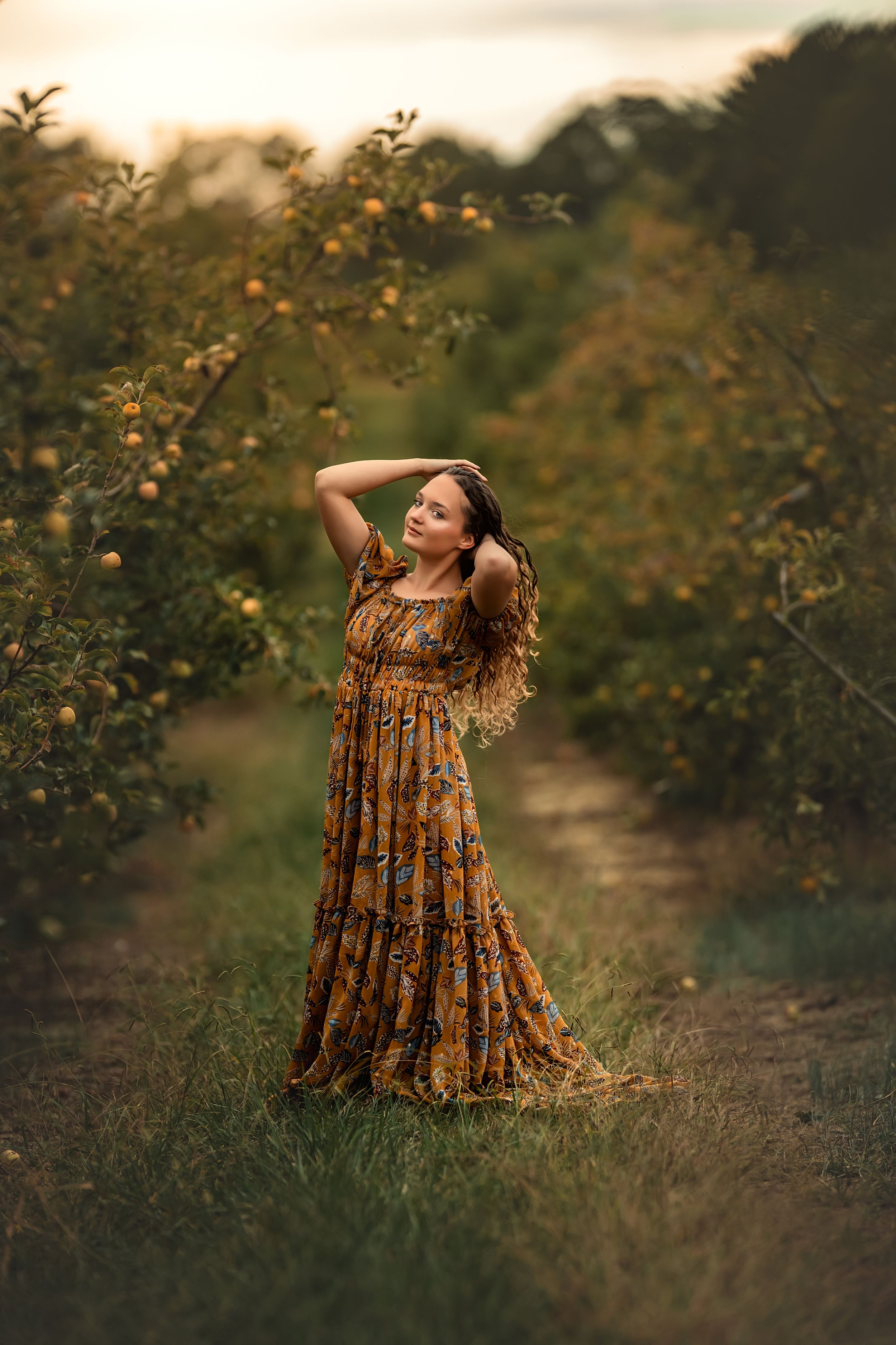 A young woman poses gracefully in an apple orchard in a golden floral dress.