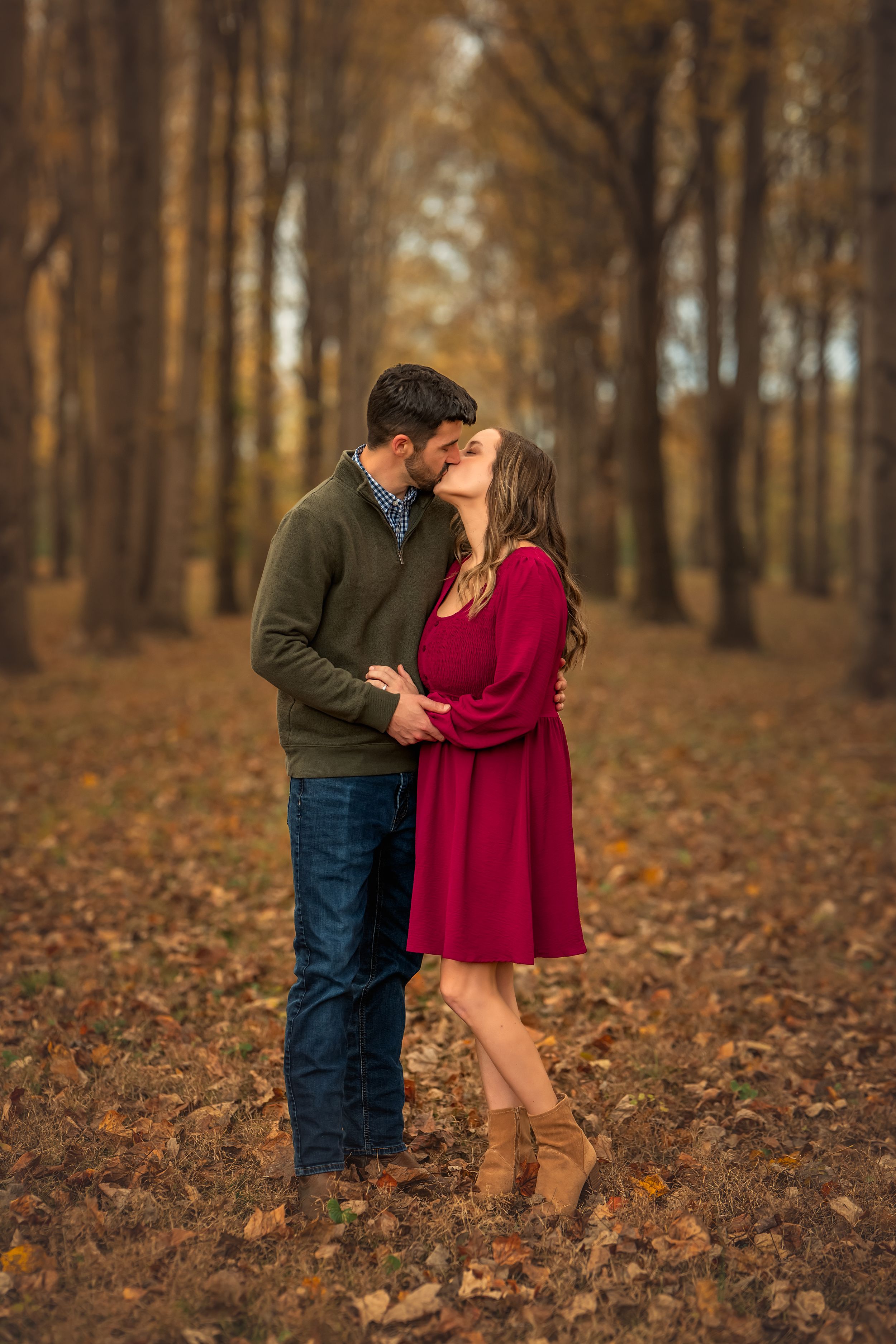 A couple embraces in a romantic autumn forest setting with fallen leaves on the ground and golden light filtering through trees.