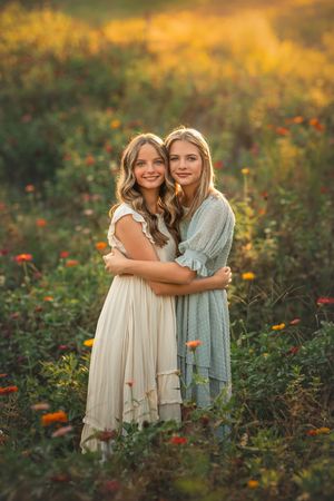 Twin sisters in flowy dresses embrace while standing in a wildflower meadow at golden hour.