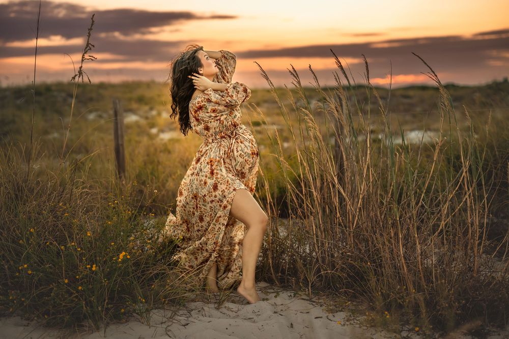 A pregnant woman in a flowy floral dress catches the wind against a golden sunset in a field of tall grass.