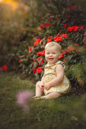 A smiling baby in yellow sitting next to vibrant red flowers in a garden setting for a family photo session with Marietta photographer.