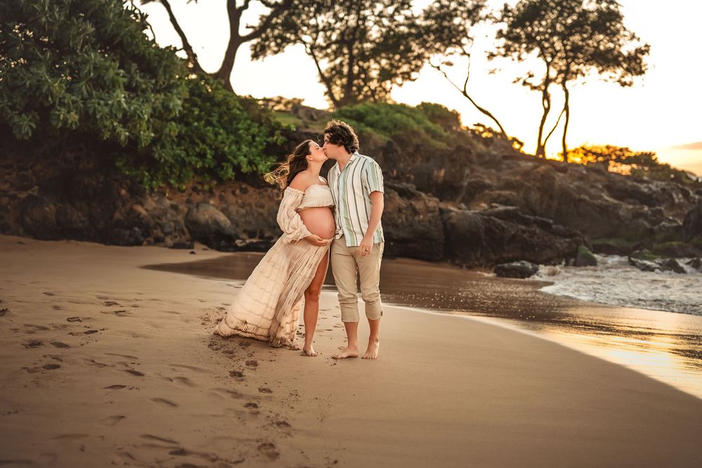 Pregnant couple embraces on a tropical beach at sunset with rocky cliffs and ocean waves in the background.