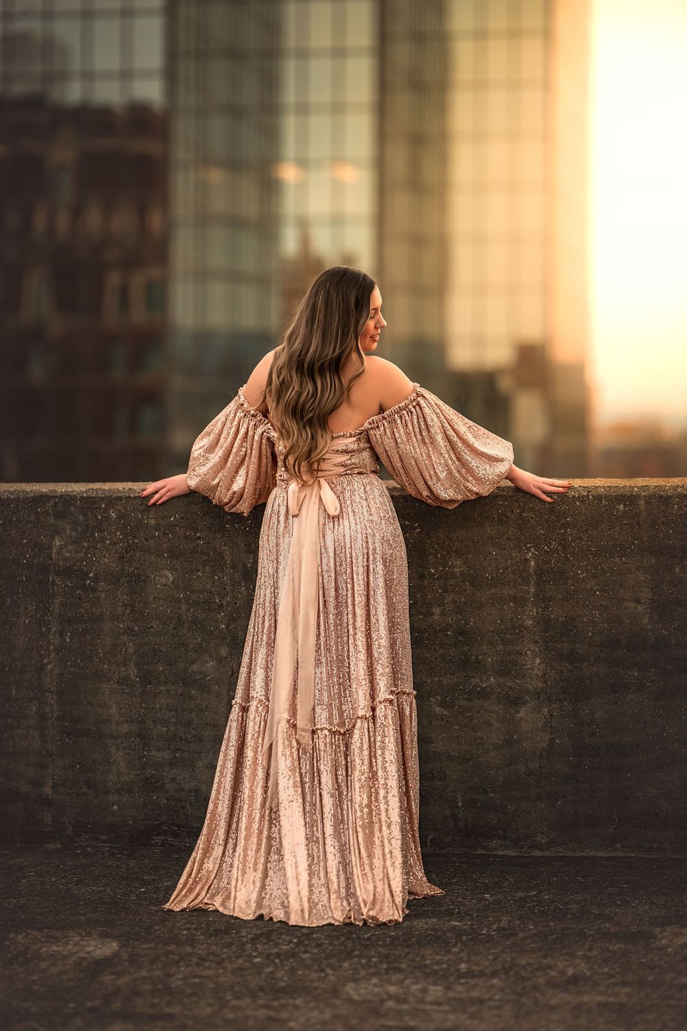A woman shows off the back of a flowing pink sequined dress that sparkles against an urban background on top of a parking garage.