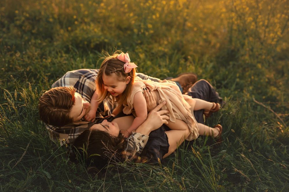Mom and Dad share a tender moment with their little girl, lying in a sunlit meadow during golden hour for a family photo session.