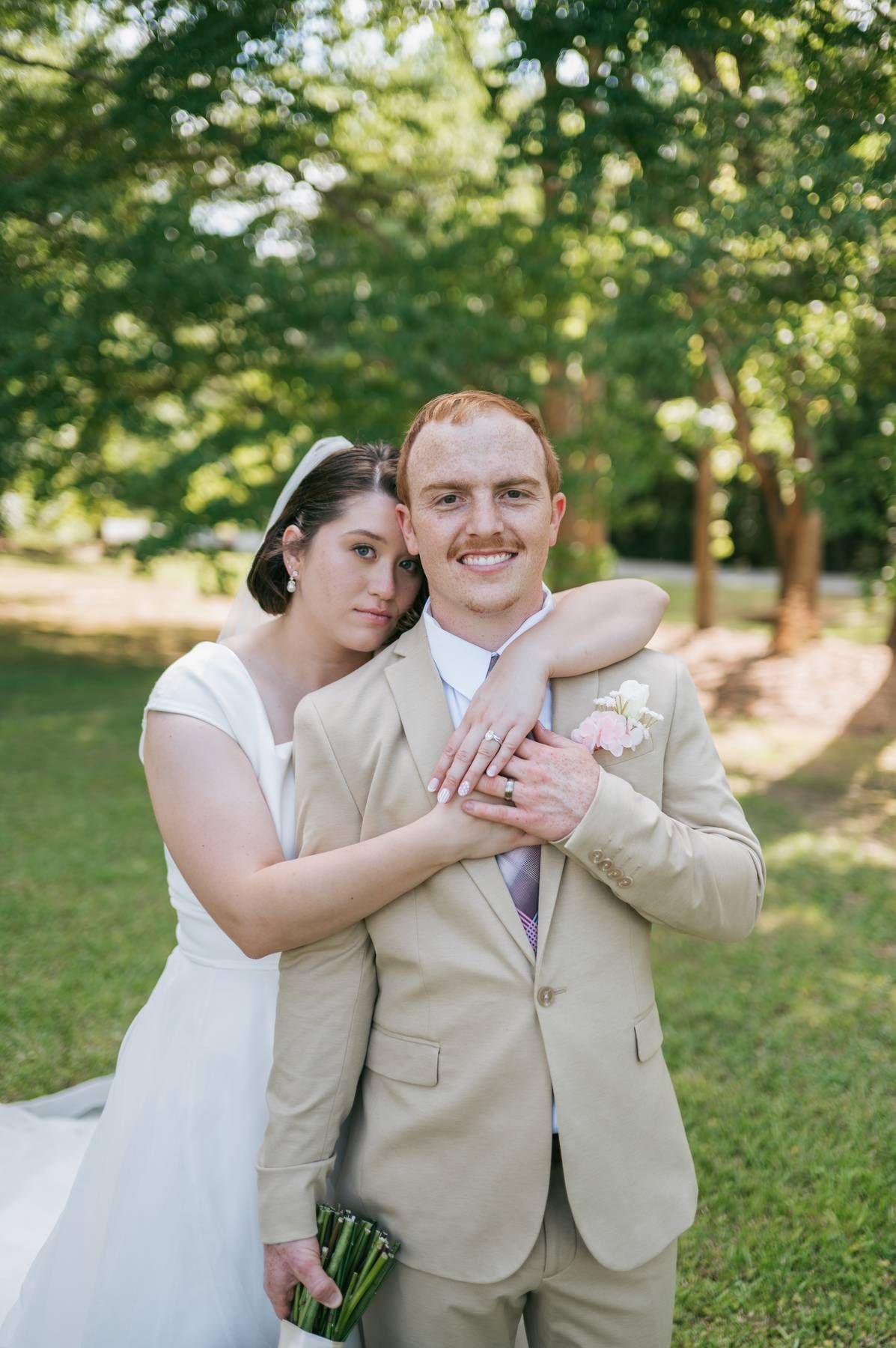 Smiling groom in tan suit embraced by bride in white dress in a sunny park setting Asheboro NC