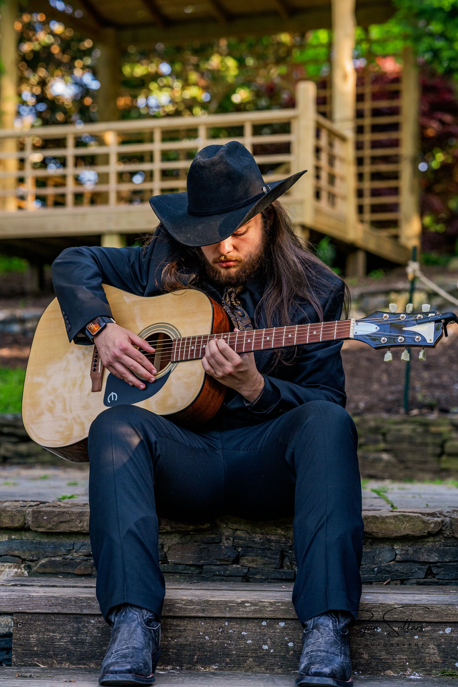 Musician in black cowboy hat playing acoustic guitar while seated on outdoor stone steps.