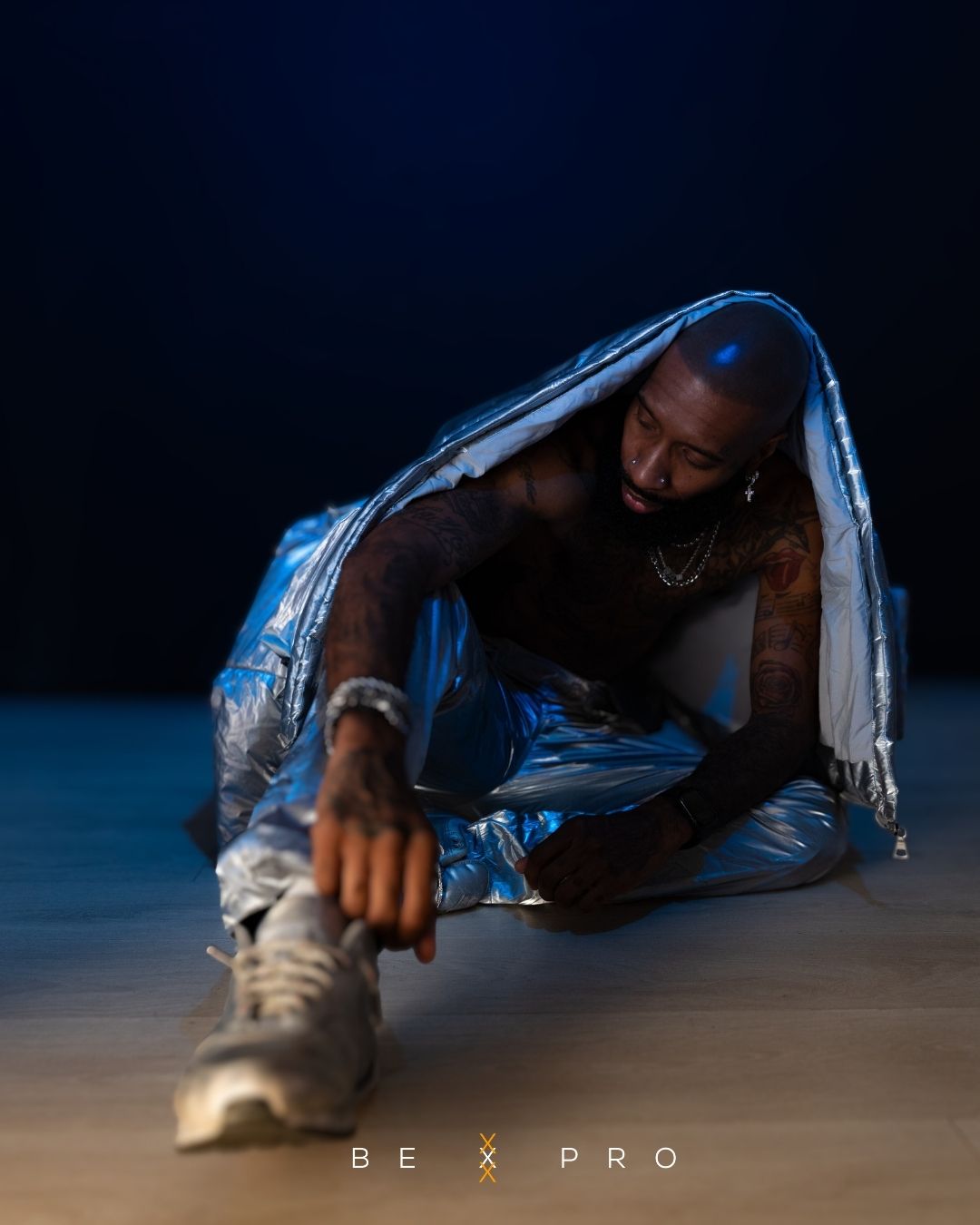 Tattooed male dancer poses dramatically on floor wrapped in silver metallic jacket under blue light.