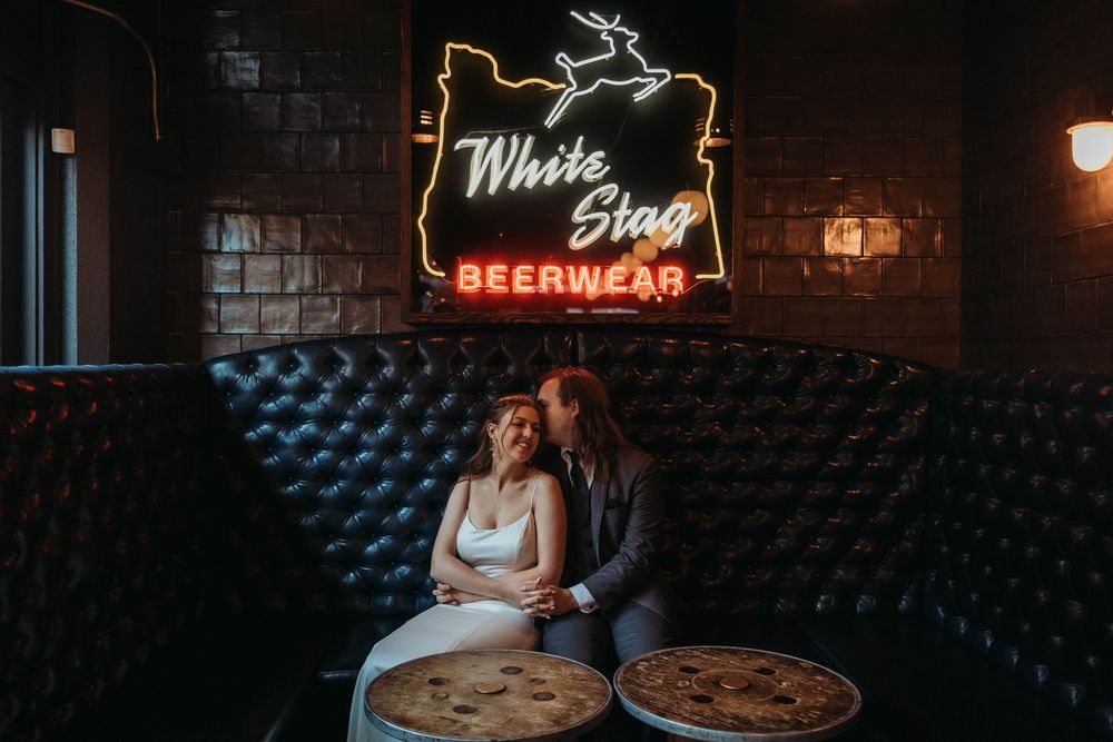 Couple sitting at table under vintage White Stag neon sign in dimly lit bar.