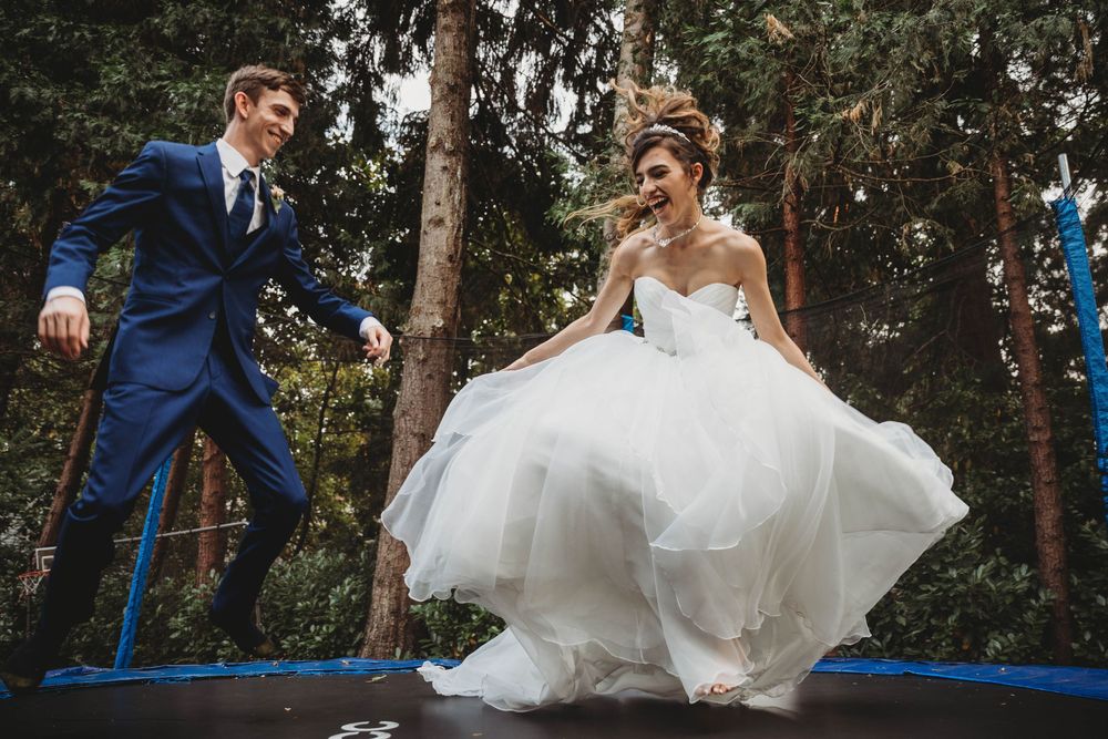 Playful newlyweds jumping on trampoline in formal wedding attire among trees.