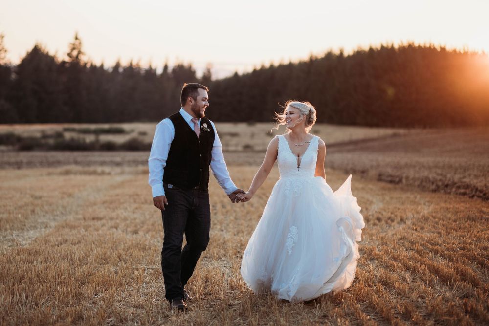 Couple walking hand in hand through golden field at sunset in wedding attire.