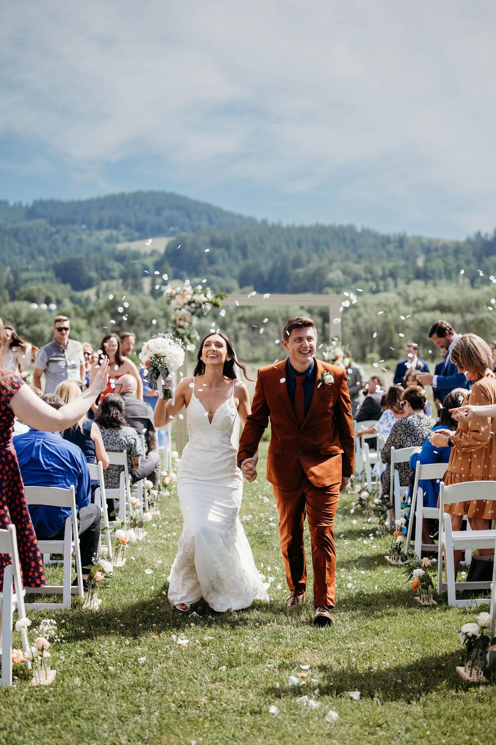 Newlyweds walk down aisle at outdoor mountain vineyard ceremony as guests cheer.