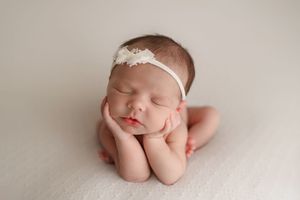 Sleeping newborn baby girl in froggy pose wearing white floral headband on cream background.