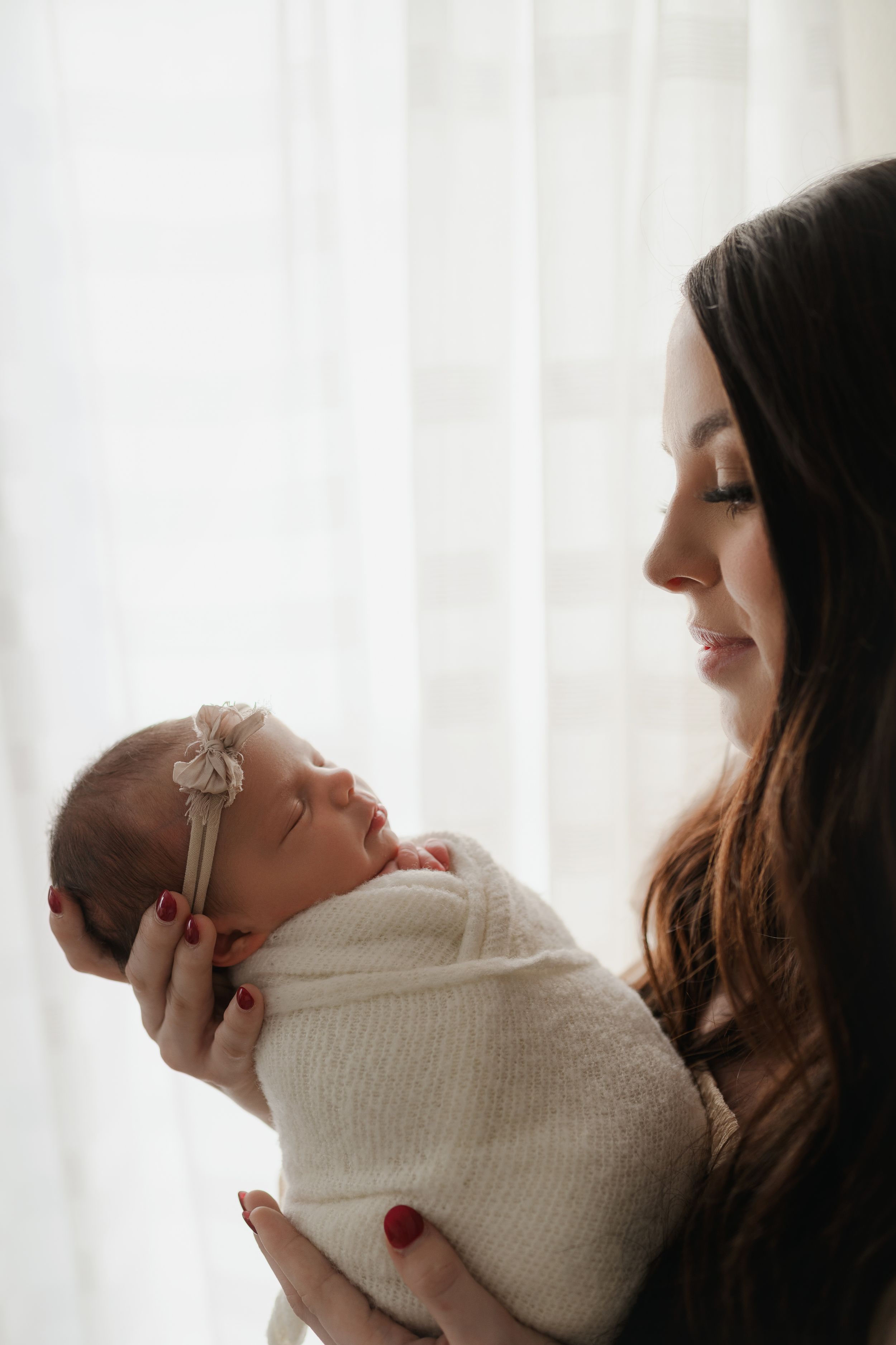 Mother tenderly gazing at swaddled newborn baby girl held in arms near bright window.