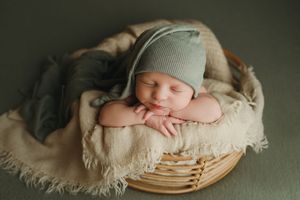 Sleeping newborn in green knit bonnet posed in wooden basket with beige blanket.
