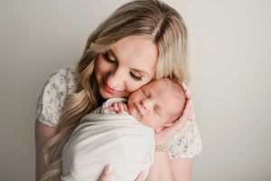 Smiling blonde mother in floral dress cuddles sleeping swaddled newborn baby close.