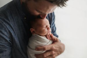 Loving father in blue shirt kisses sleeping swaddled newborn baby on forehead.